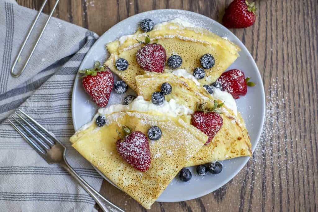 overhead photo of three sourdough crepes on a gray plate and topped with whipped cream, blueberries, strawberries and powdered sugar