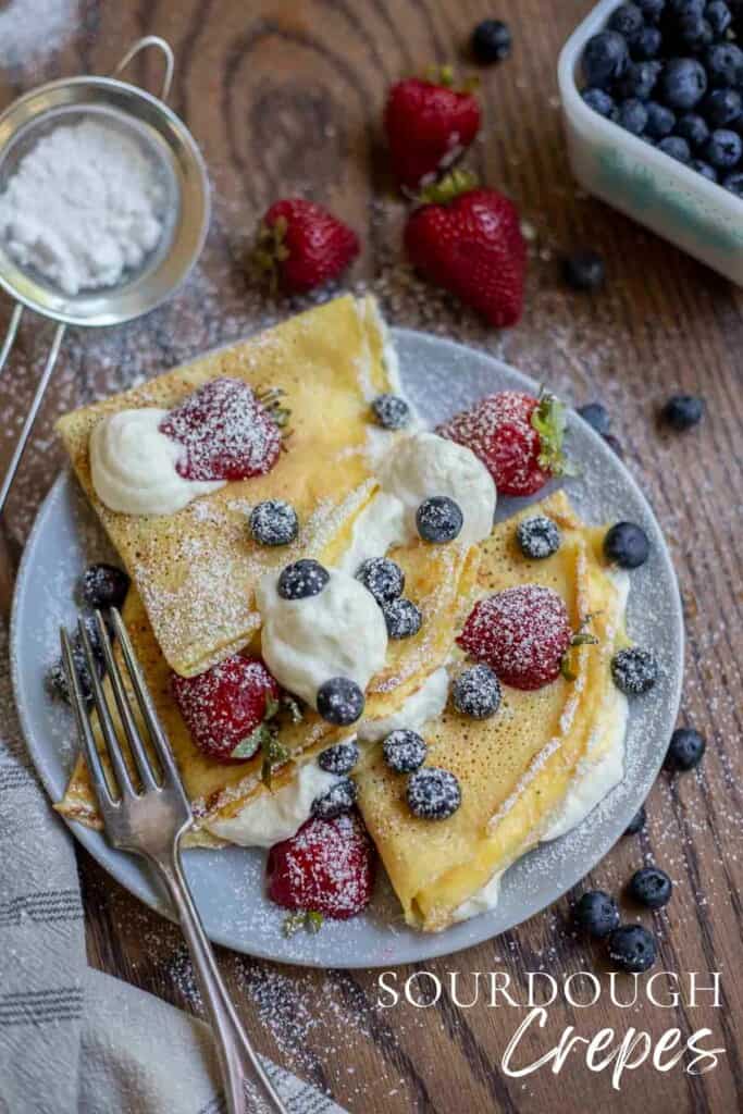 three sourdough crepes with whipped cream filling and topped with blueberries, strawberries and powdered sugar on a gray plate on a wood table with ingredients spread around the plate