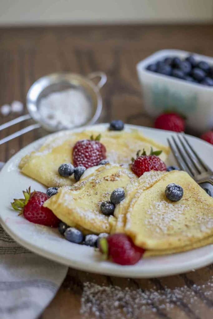three sourdough crepes topped with blueberries and strawberries on a white plate with a dish of blueberries and a sifter of powder sugar in the background