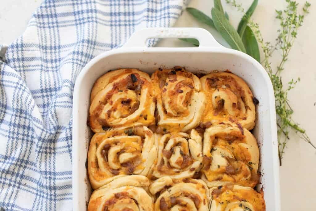overhead photo of half a baking dish full of sourdough cheese and herb rolls on a blue and white plaid towel with fresh herbs to the right of the pan