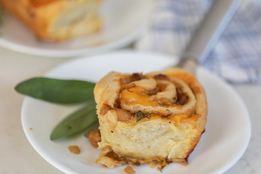 sourdough herb and cheese roll on a white plate with sage on a white countertop with a blue and white towel in the background