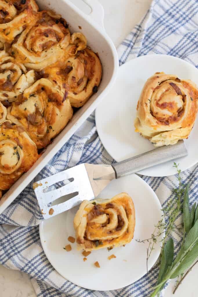 overhead photo of sourdough herb and cheese rolls in a baking dish with two plate with rolls to the right on a top of a blue and white plaid towel