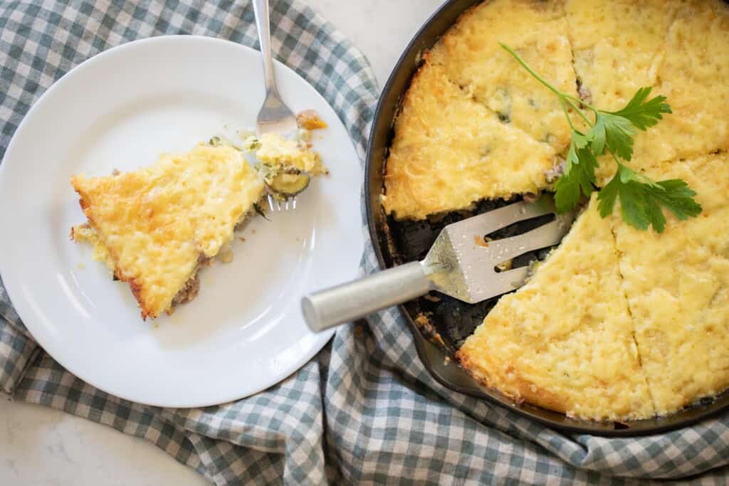 sourdough skillet topped with cheese and parsley in a cast iron skillet with a slice cut out. The slice is on a white plate on a blue checked towel