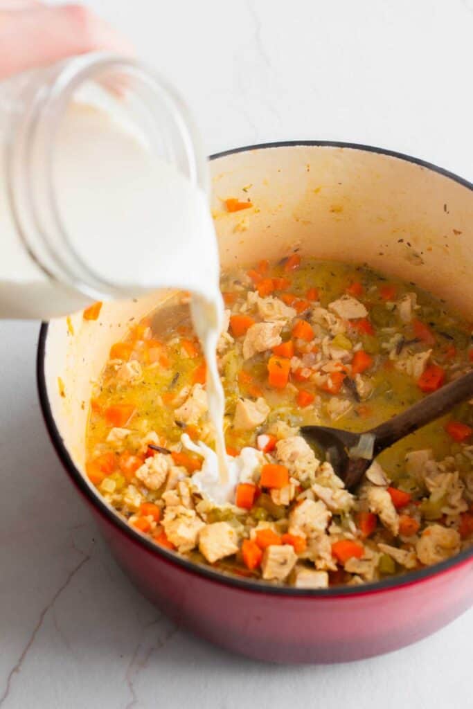 A dutch oven with soup with a mason jar of cream being poured into it. 