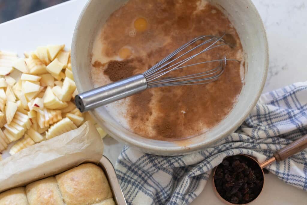 large bowl with eggs, milk, sugar, and spices. chopped apples and a loaf of bread to the left and a towel and measuring cup of raisin to the right