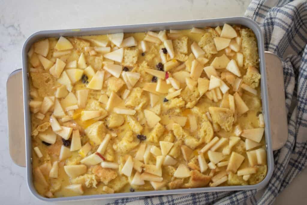 custard poured over cubed bread, chopped apples and raisins in a baking dish