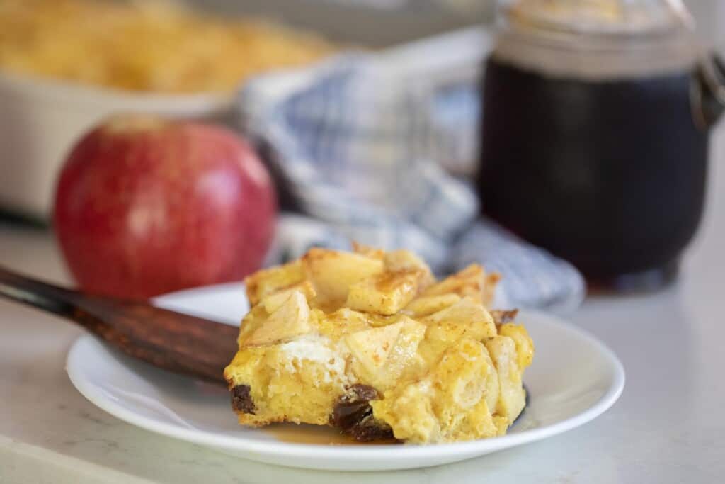 slice of sourdough French toast casserole on a white plate with a wooden spatula. A apple, blue plaid towel and a jar of maple syrup is in the background