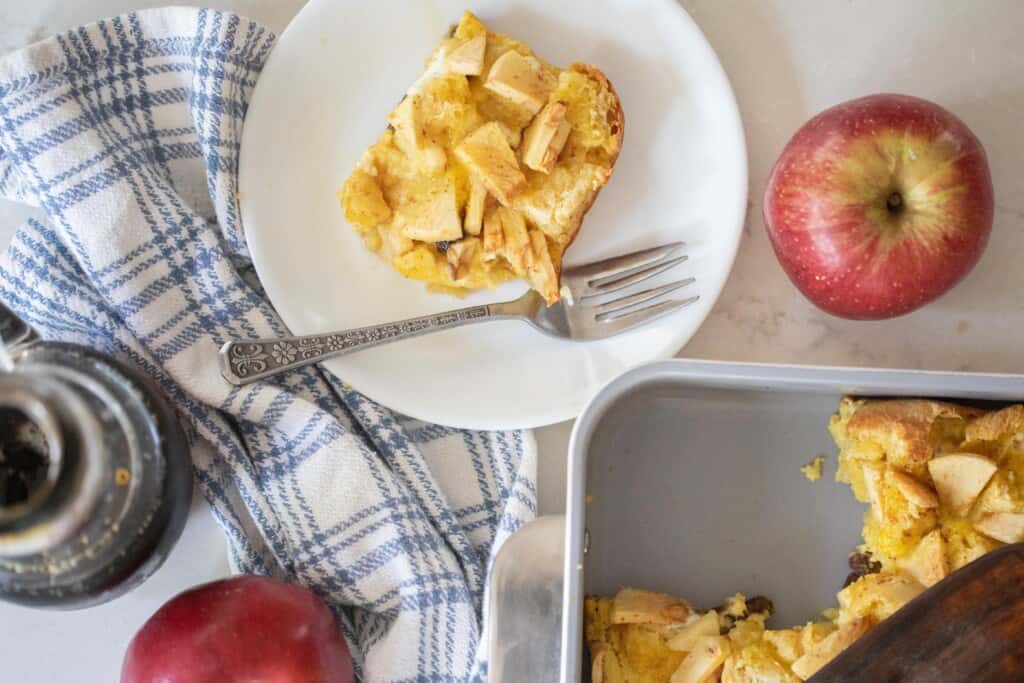 overhead photo of French toast casserole with apples and raisins on a white plate with a fork. An apple sits on the counter to the right along with a baking dish of more casserole.