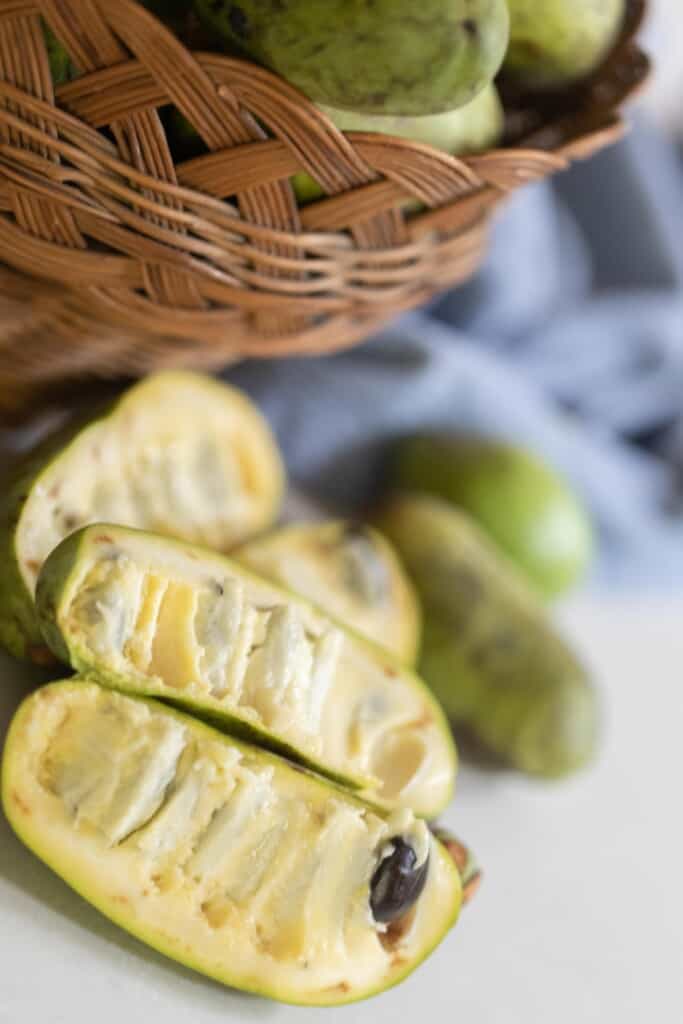 pawpaws sliced in half on a white countertop with a baskets of pawpaws and a blue towel in the background