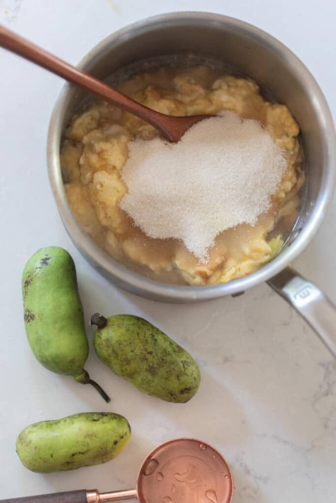 sugar and pawpaw pulp in a saucepan with a wooden spoon on a white countertop. A copped measuring scoop and 3 fresh pawpaws is it on the counter in front of the pot