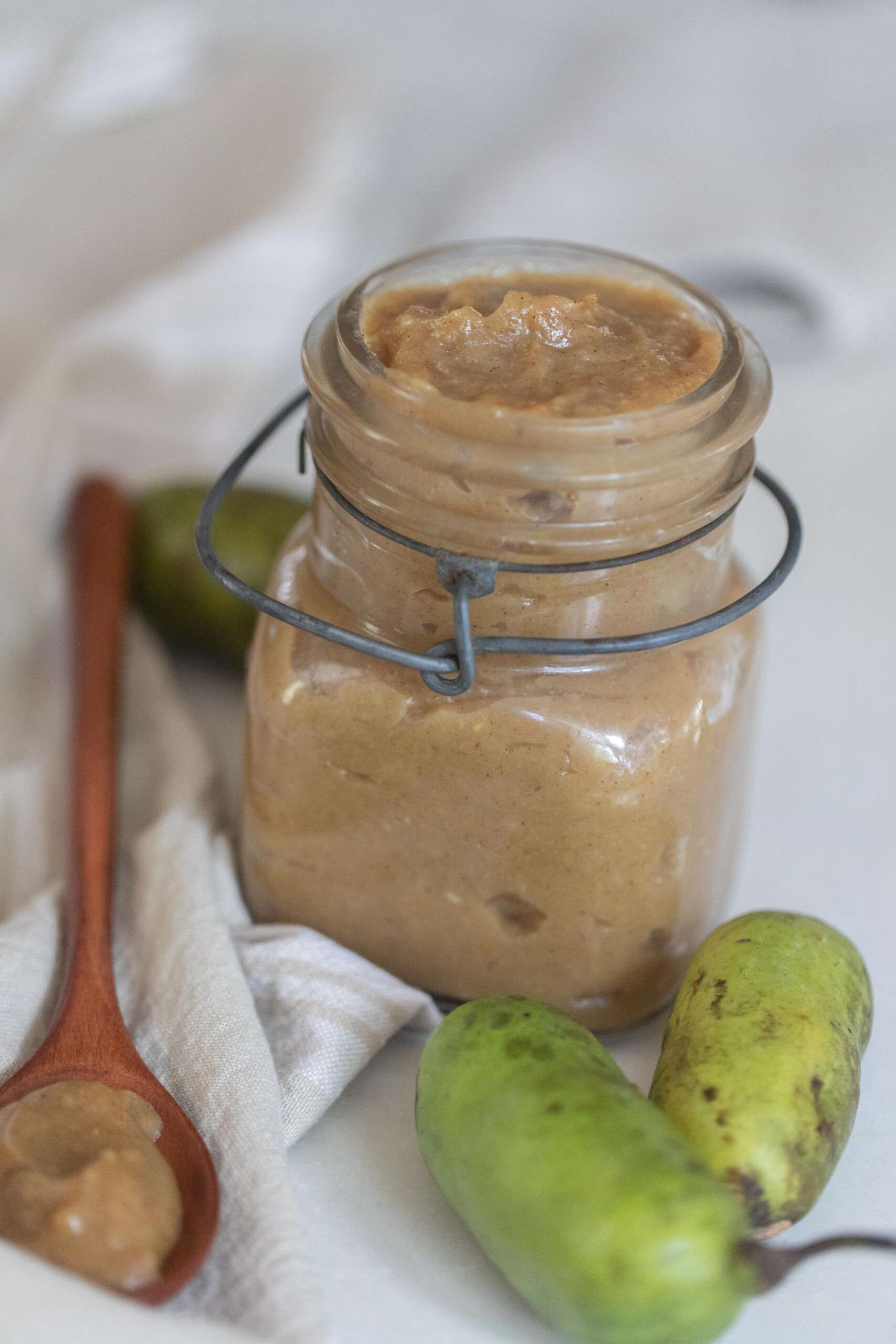 a swing top mason jar with pawpaw jam on a white towel with fresh pawpaw and a wooden spoons with jam in front of the jar