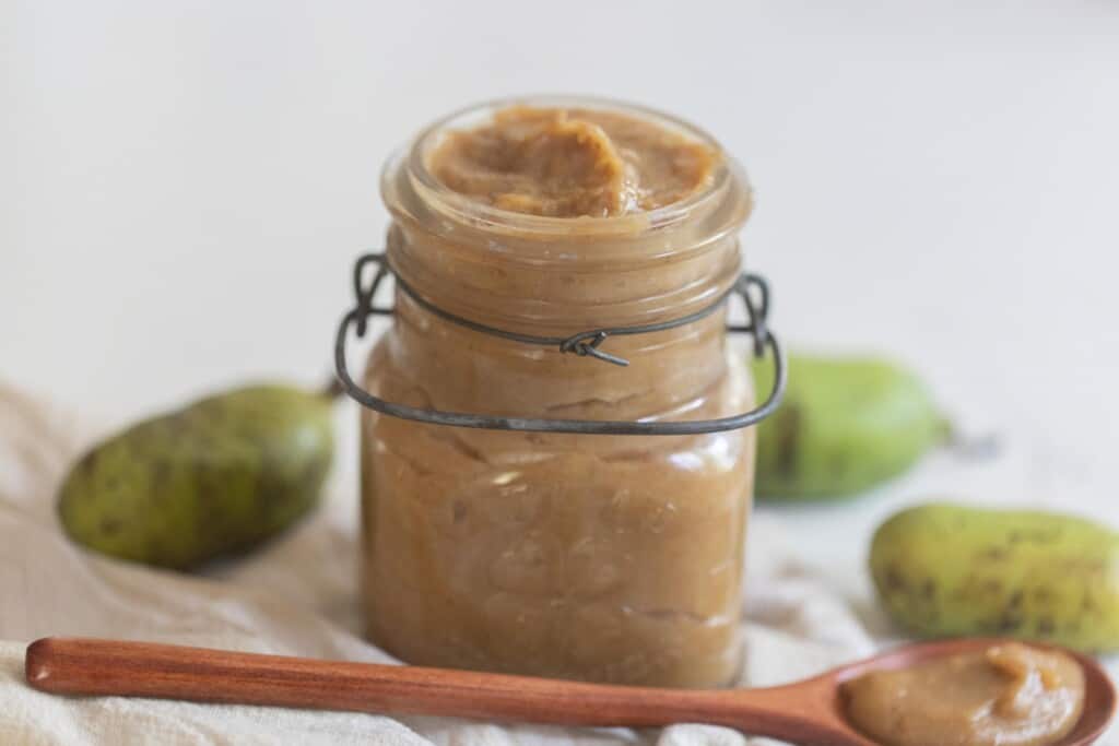 pawpaw jam in a glass swing top jar with pawpaws in the background and a wooden spoon in front of the jar