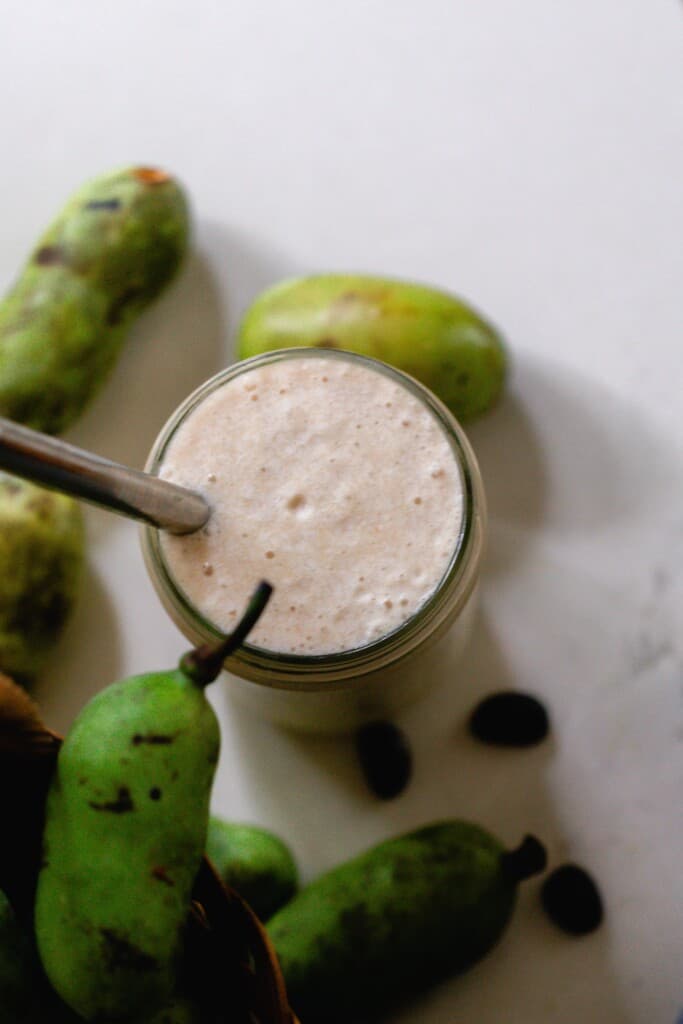 overhead photo of a pawpaw smoothie in a glass jar with a straw on a white countertop. Fresh pawpaws surround the jar