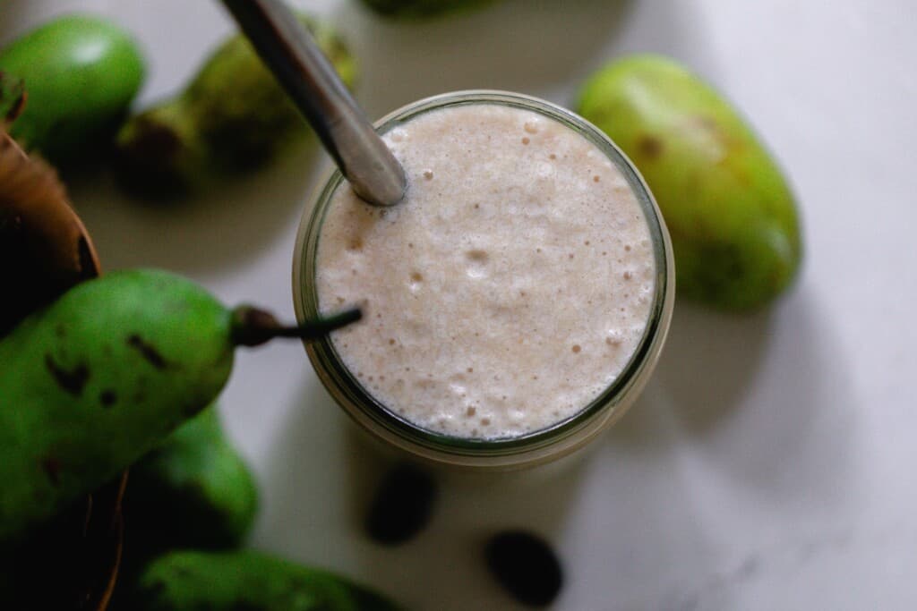 overhead photo of a pawpaw smoothie with a metal straw in a mason jar. The jar sits on a white countertop surrounded by fresh pawpaws