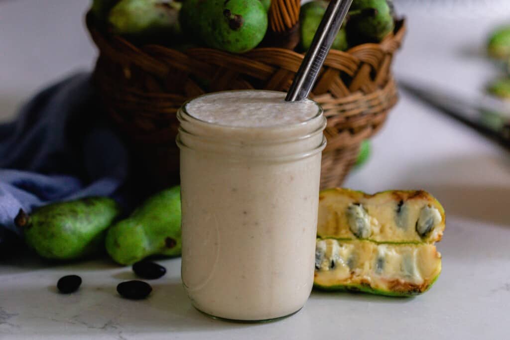 pawpaw and banana smoothie in a mason jar with a metal straw. The smoothie is on a white countertop with fresh pawpaws on a basket in the background