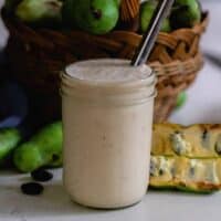 pawpaw and banana smoothie in a mason jar with a metal straw. The smoothie is on a white countertop with fresh pawpaws on a basket in the background