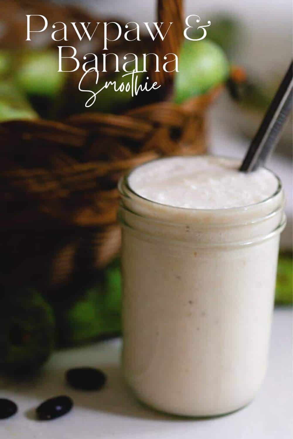 a creamy pawpaw smoothie in a glass mason jar with a metal straw on a white countertop with a basket of pawpaws in the background