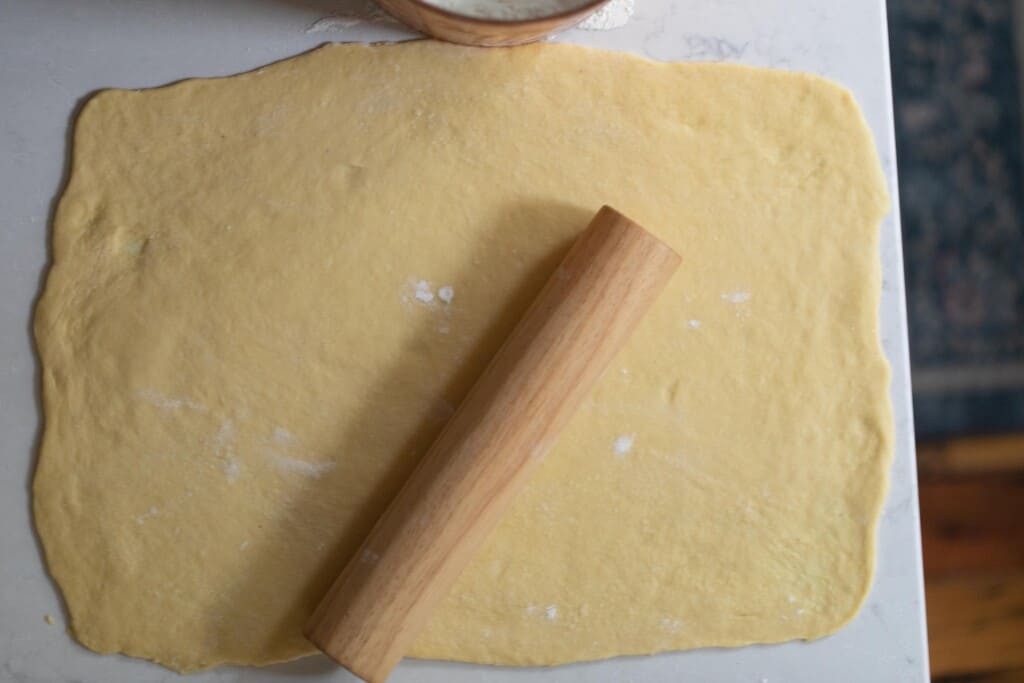 noodle dough rolled out on a white countertop with a wood rolling pin resting on top