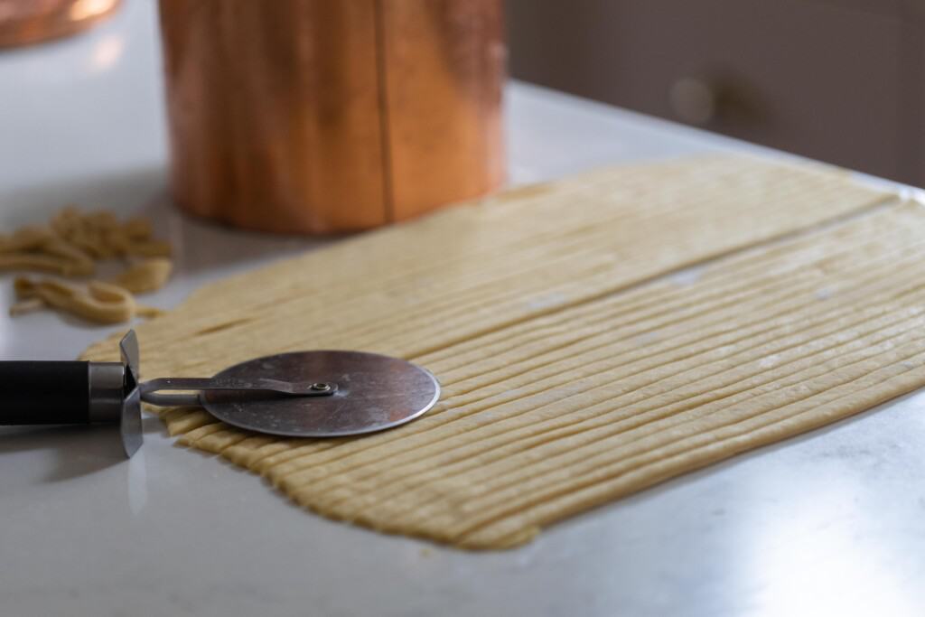 noodle dough rolled out onto a white countertop and sliced with a pizza cutter. A pizza cutter sits on the dough with a copper canister in the background 