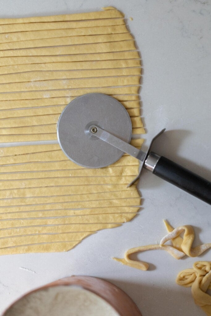 homemade noodles rolled out onto a rectangle on a white countertop sliced with a pizza cutter. The pizza cutter is sit-in ton the dough
