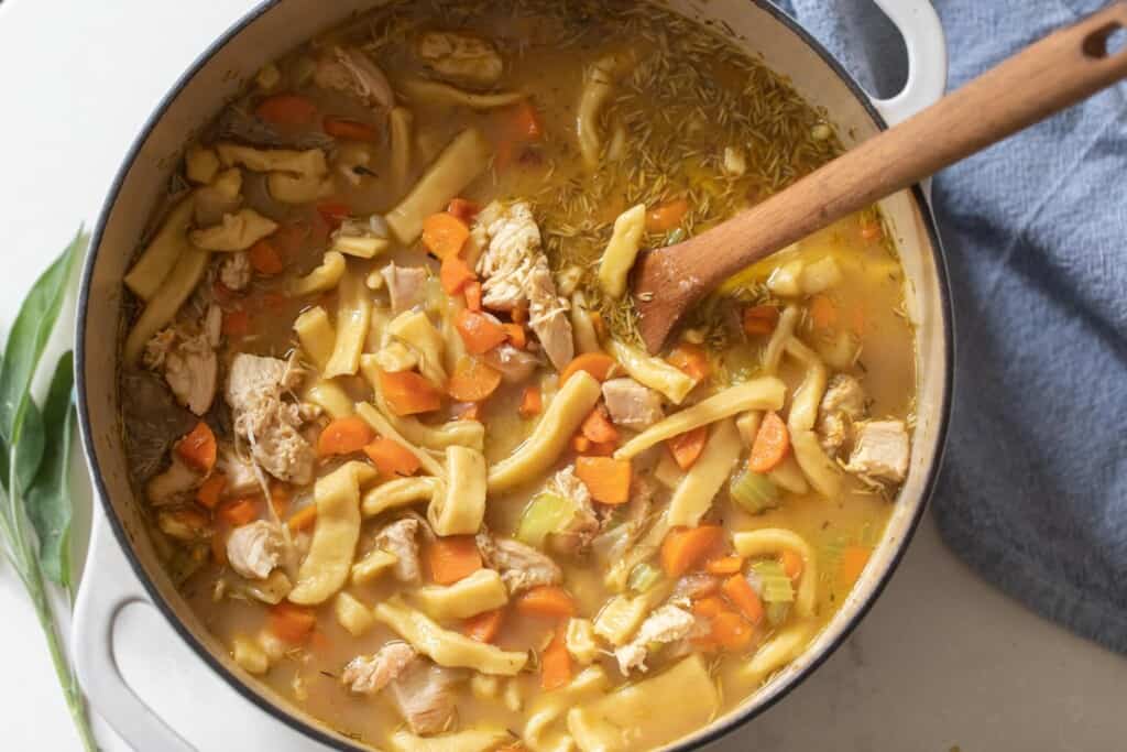 overhead photo of a white dutch oven full of homestyle chicken noodle soup with carrots with a wooden ladle in the pot a blue pot is to the right