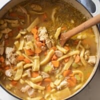 overhead photo of a white dutch oven full of homestyle chicken noodle soup with carrots with a wooden ladle in the pot a blue pot is to the right