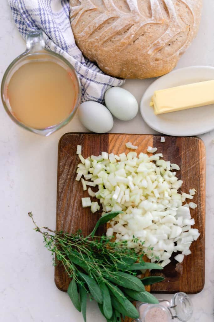 sourdough bread, eggs, broth, butter on a white countertop. A wood cutting board with celery, onions, and fresh herbs