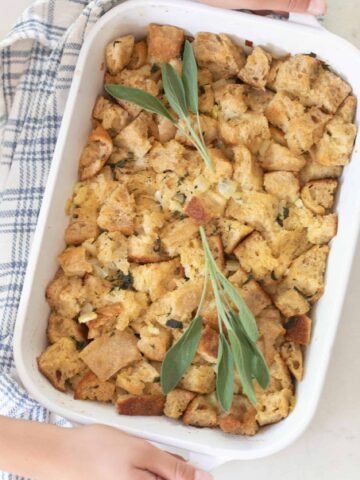 hands holding a white casserole dish full of homemade sourdough stuffing topped with fresh sage