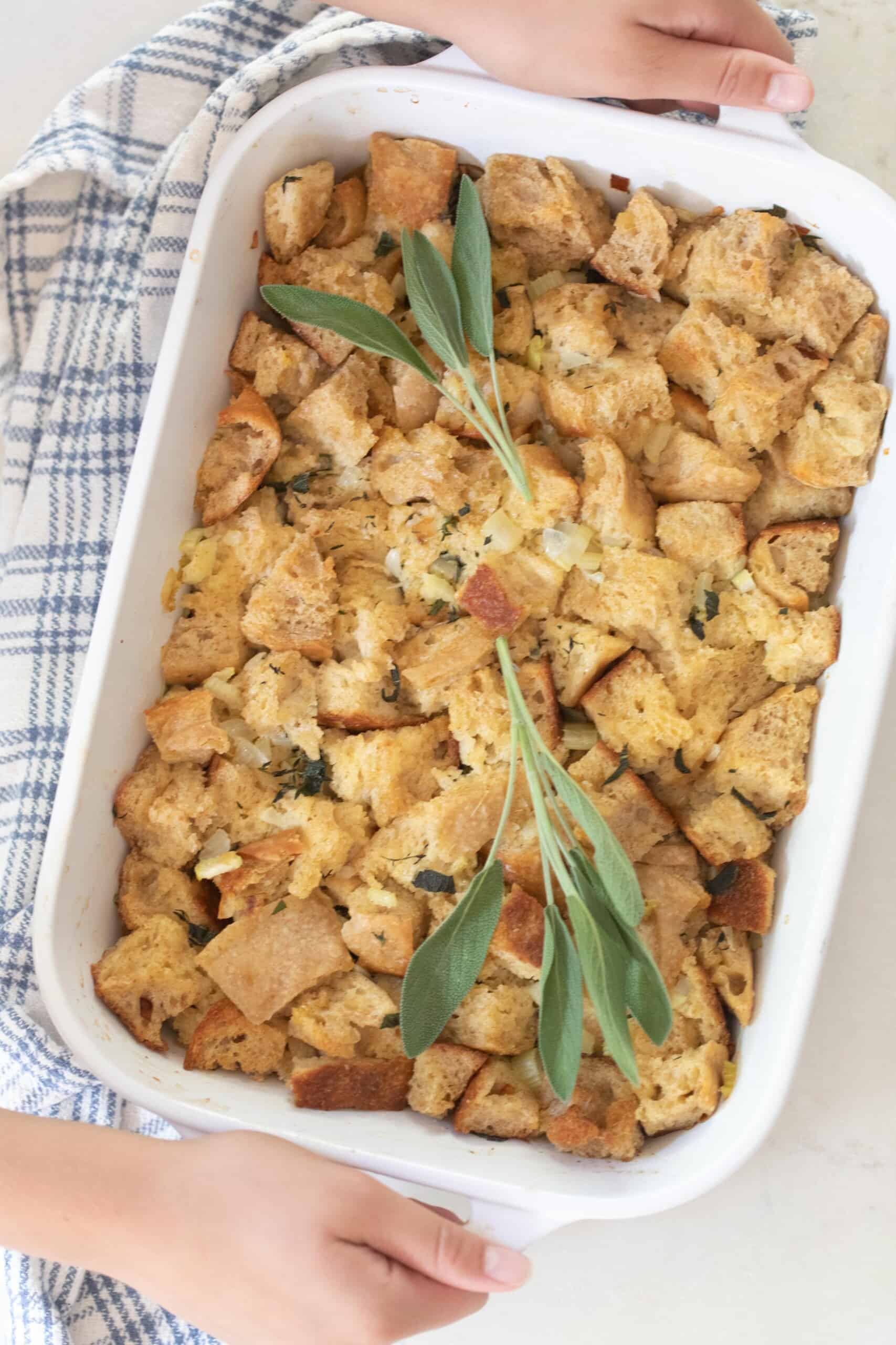 hands holding a white casserole dish full of homemade sourdough stuffing topped with fresh sage