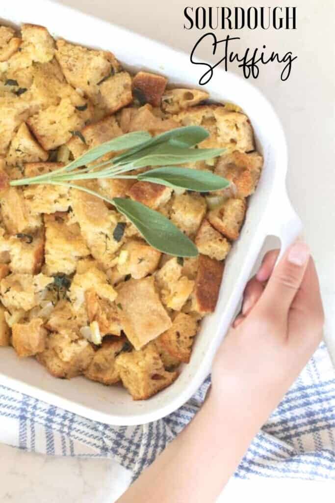 overhead photo of a hand holding a baking dish rim on a white countertop. The baking dish has sourdough stuffing topped with fresh sage