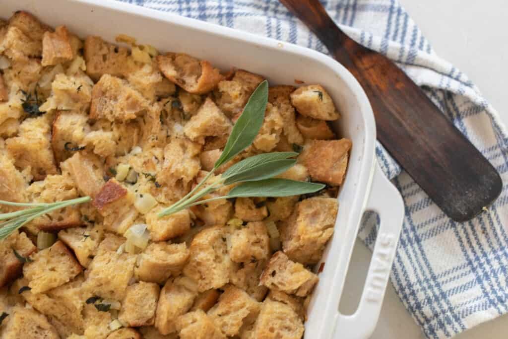sourdough stuffing topped with fresh sage in a white baking dish on a white and blue plaid towel with a wood spatula resting on top of the towel