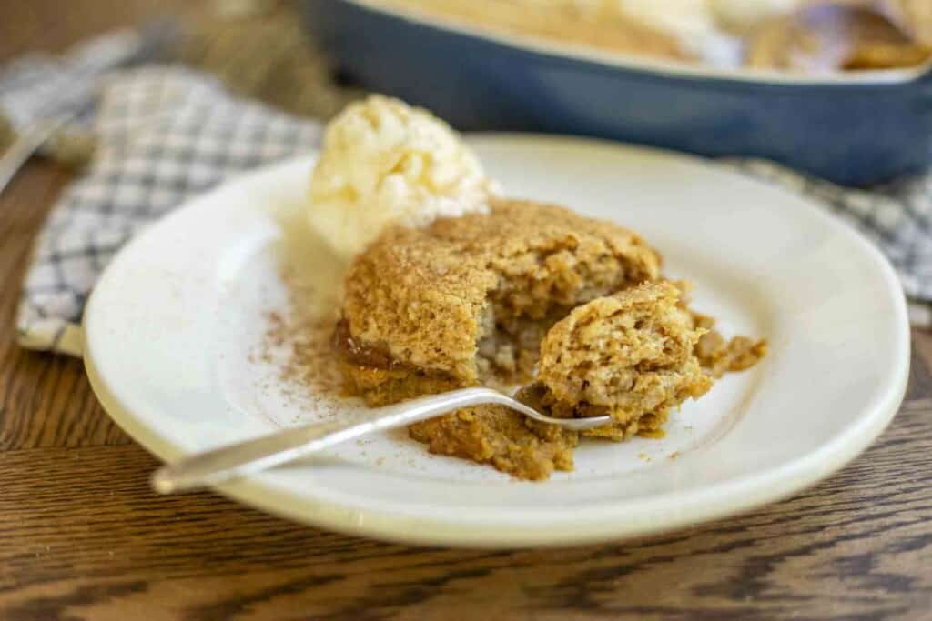 plate of sourdough pumpkin cobbler on a white plate with a spoon with a spoonful of cobbler and ice cream on the plate. A blue baking dish is in the background on a white and black towel