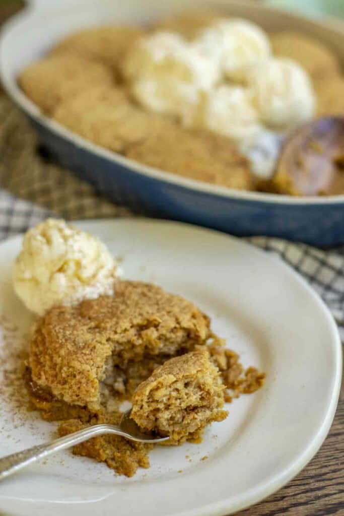 a white plate with sourdough pumpkin cobbler with a spoon with cobbler to the right. A scoop of ice cream is in the back of the cobbler. A baking dish with cobbler and ice cream in the background