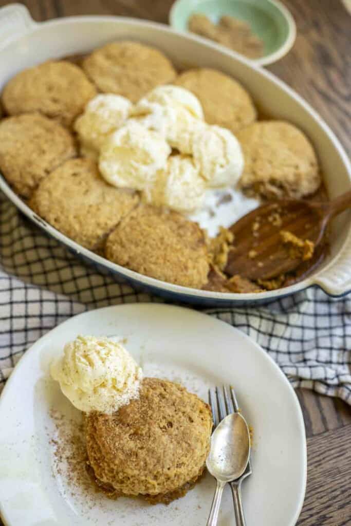 sourdough pumpkin cobbler topped with a scoop of ice cream and a spoon on a white plate. A blue baking dish with more cobbler and ice cream in the background