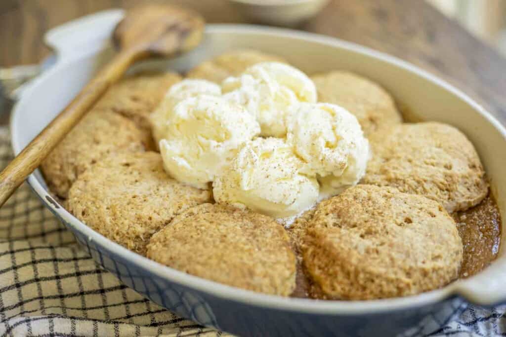 baking dish with pumpkin custard topped with a sourdough biscuit topping topped with vanilla ice cream in a blue baking dish. A wooden spoon is resting on top of the baking dish 