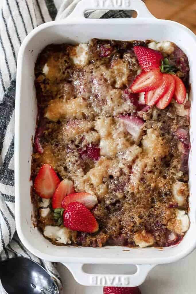 overhead photo of strawberry cream cheese sourdough cobbler in a white baking dish topped with strawberries on a table with a white and black towel to the left