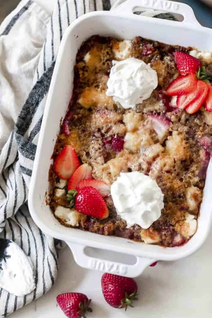 sourdough strawberry cobbler topped with fresh whipped cream and sliced strawberries. The cobbler is in a white baking dish on a white and black stripped towel