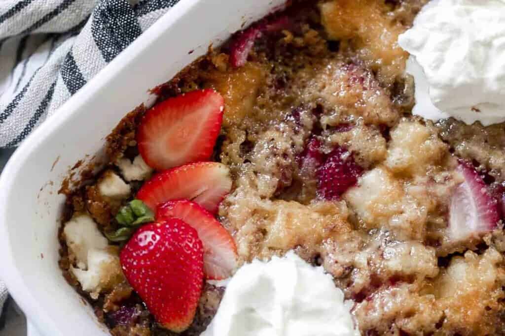 overhead photo of sourdough strawberry cobbler in a white baking dish topped with fresh strawberries and whipped cream. Only a corner of the dish is in the photo