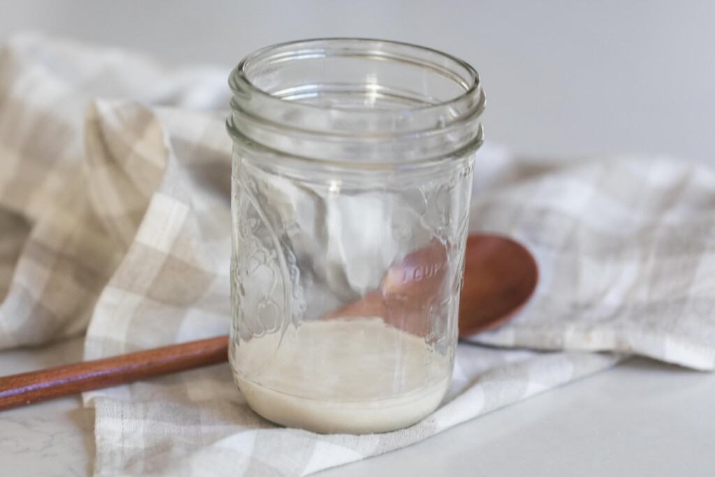 thawed sourdough starter in a glass mason jar on a white and tan checked towel with a spoon in the background