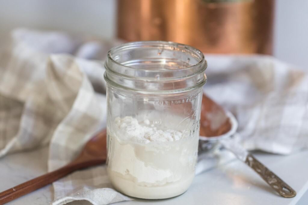 flour, water, and sourdough starter in a mason jar  on a white countertop with a wooden spoon and checked towel in the background