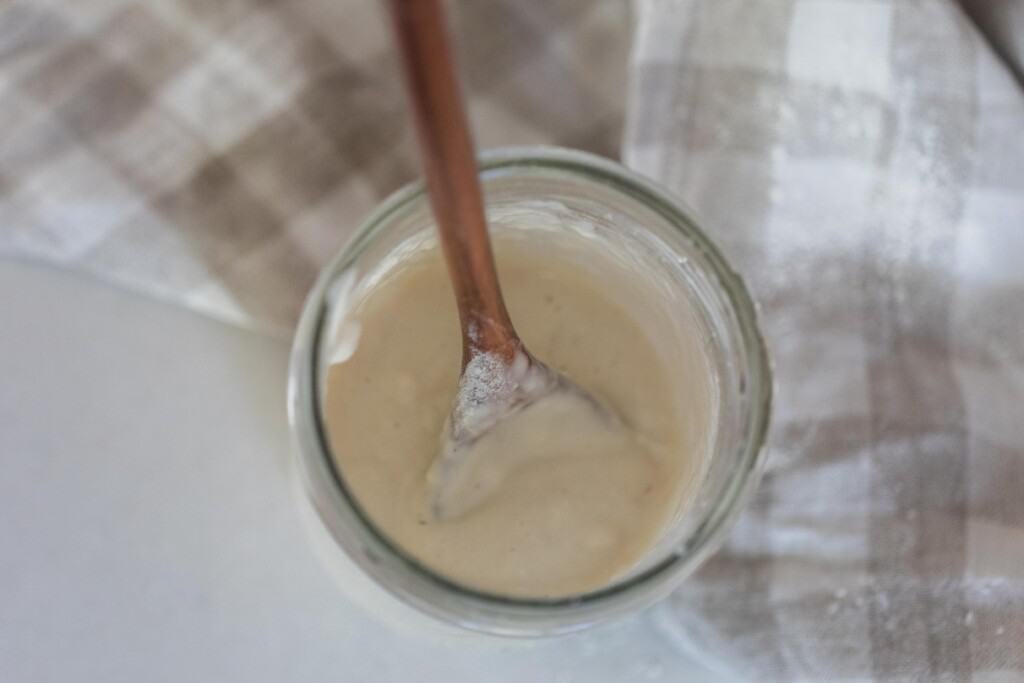 overhead photo of a sourdough starter in a mason jar with a wooden spoon in the starter, The jar sits on a tan and white checked towel