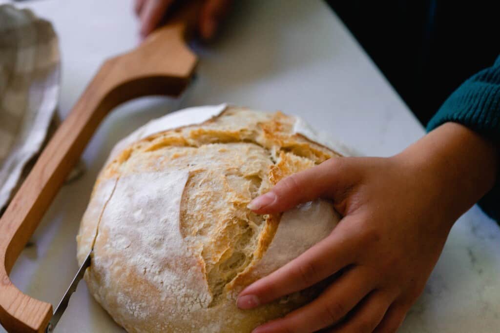 someone cutting a sourdough boule with a bow knife on a white countertop