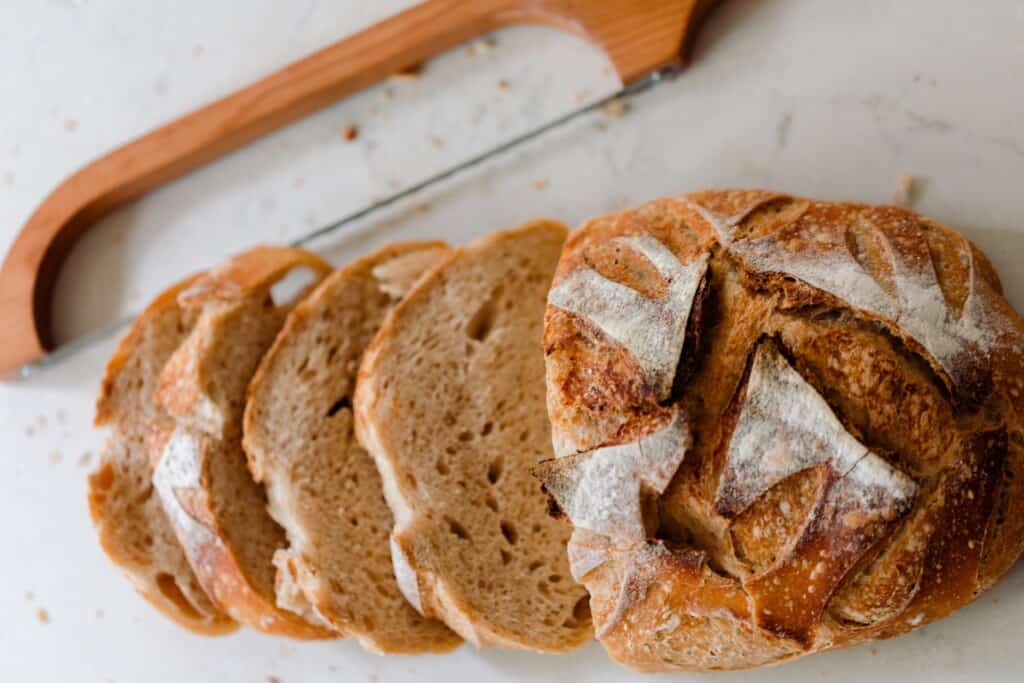 A sourdough boule cut into slices on a white countertop with a bow knife to the side