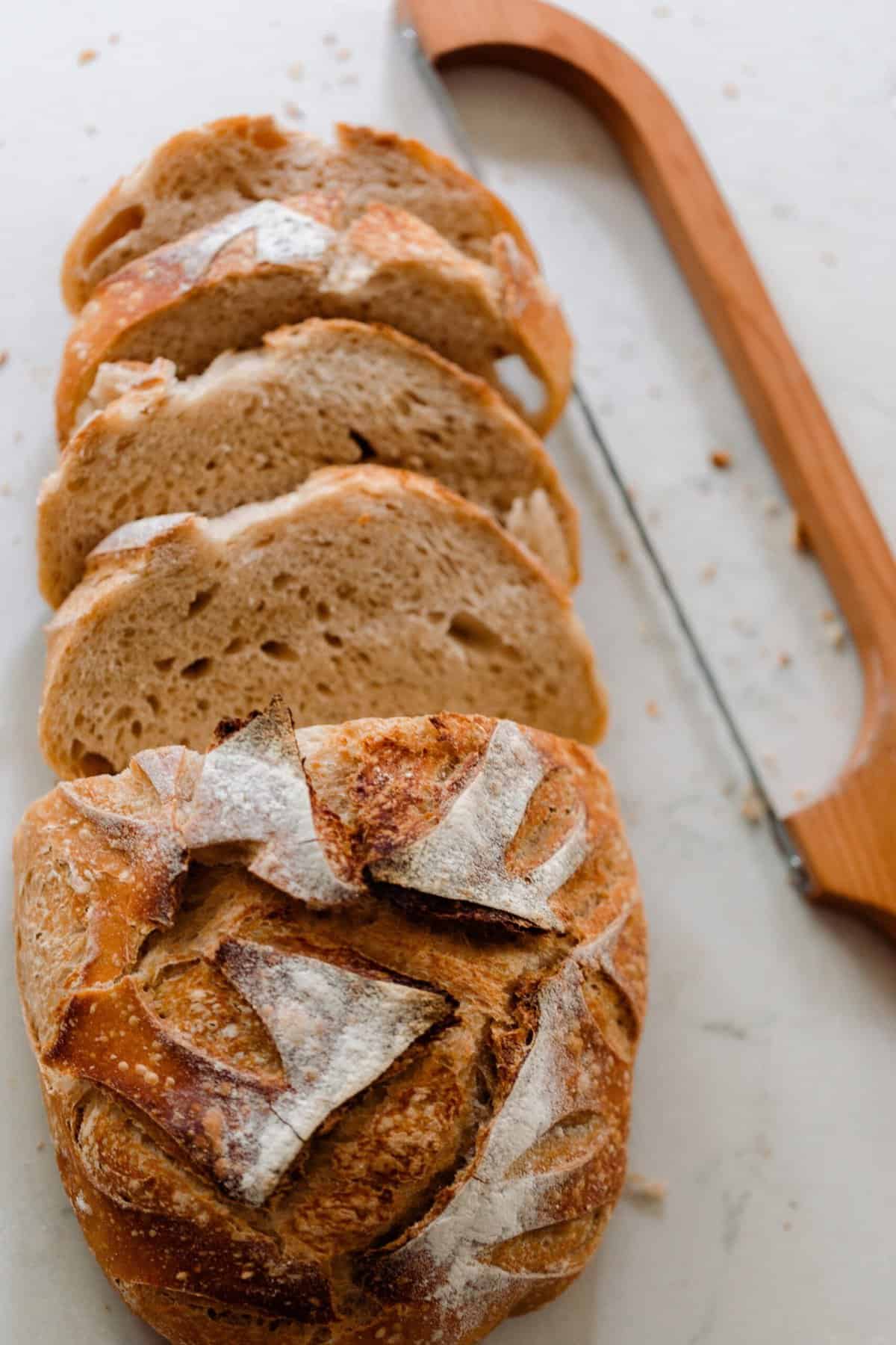 A sourdough boule cut into slices on a white countertop with a bow knife to the side