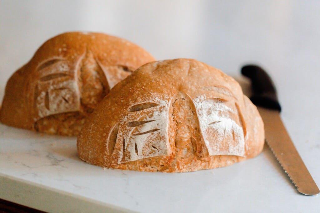 A boule of sourdough bread cut in half on a white countertop with a serrated knife on the side
