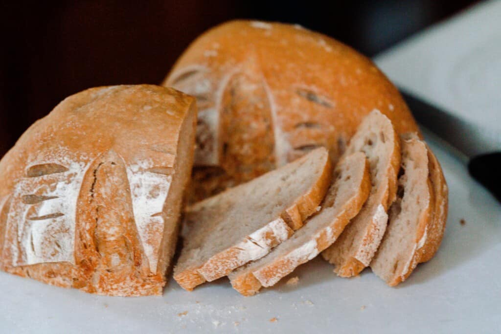 A sourdough boule cut into two halves with one cut into slices on white countertop with knife in background