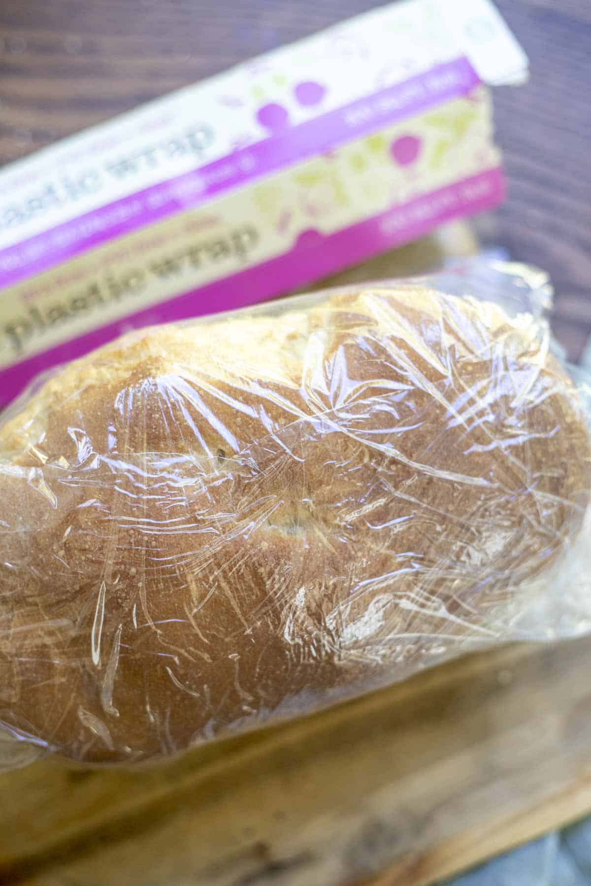 wrapping a loaf of bread in plastic wrap on a wood cutting board