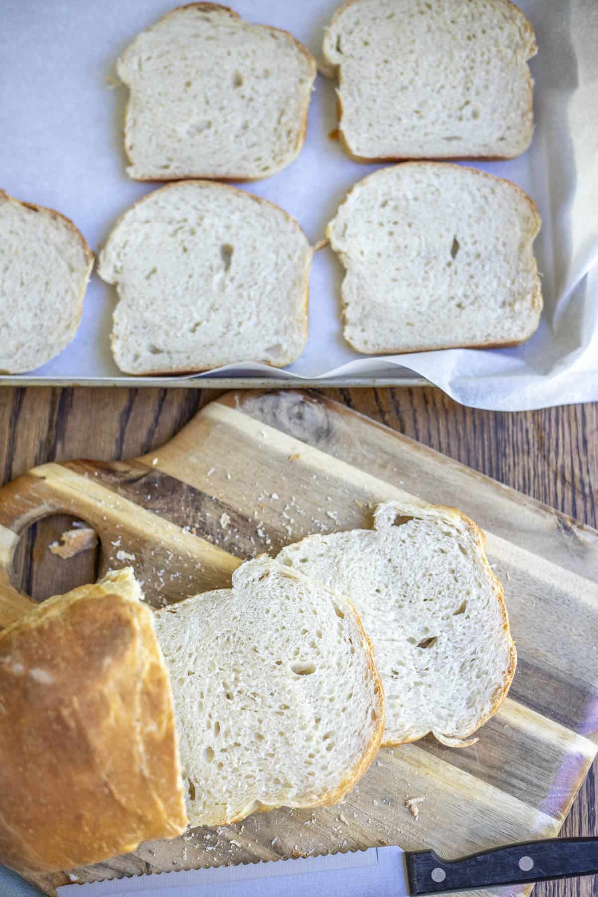 sourdough bread loaf cut on a cutting gbaord with a baking sheet of sliced bread in the background