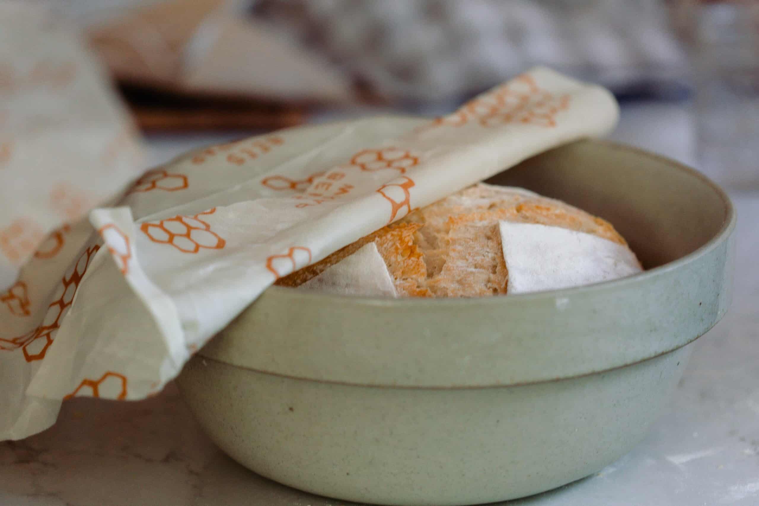 A sourdough loaf in a green bowl with a white and orange bees wax wrap covering it half way