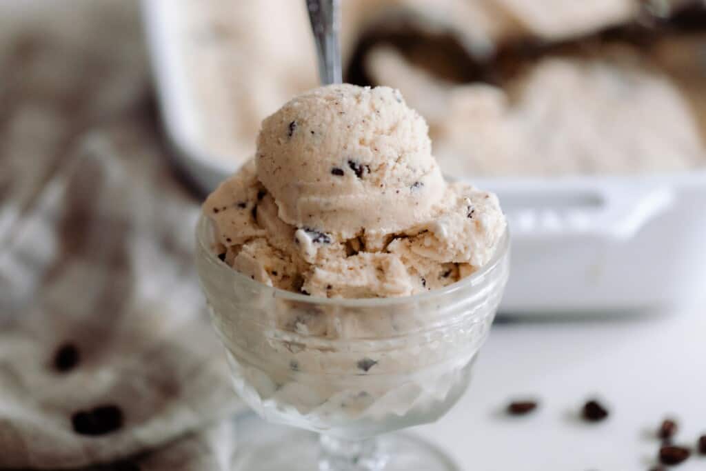 java chip ice cream in a vintage ice cream dish on a white countertop with chocolate chips, a towel and a baking dish of more ice cream in the background